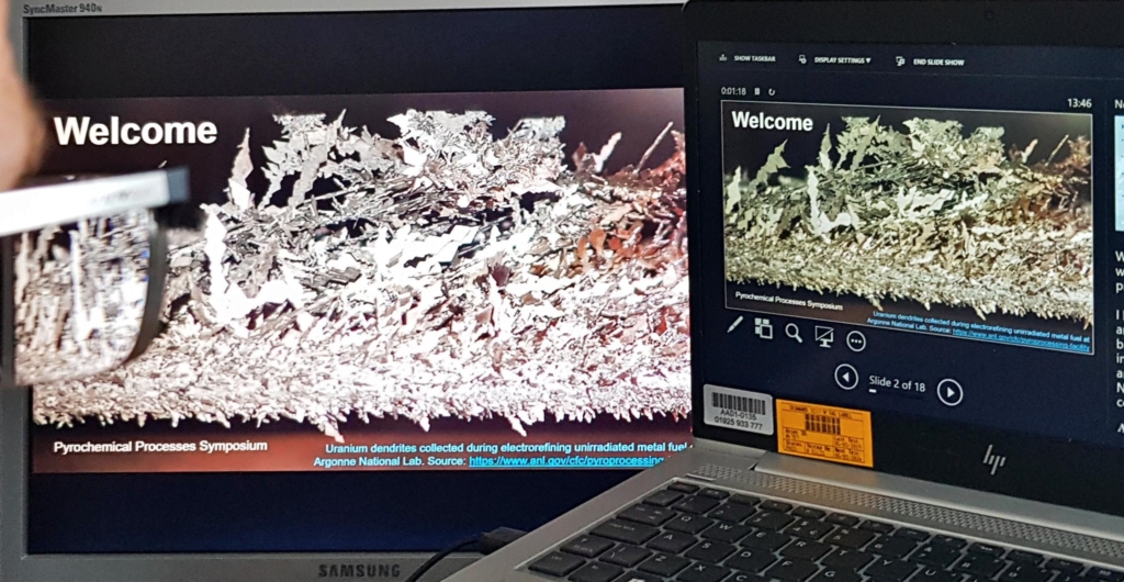 Photograph of two computer comintors showing introduction slides to the digital pyro-processing symposium. In the corner of the photo, Mike Edmondson's glasses are visible as he sits facing the screens.