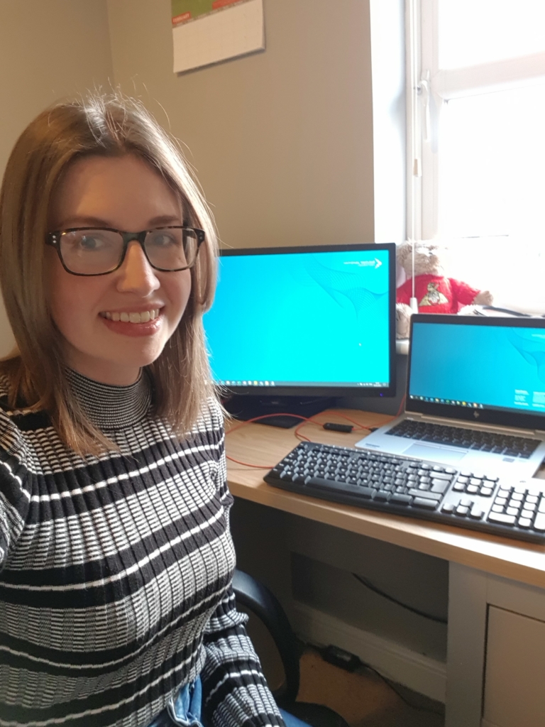 Photograph of Beth Slingsby at work, as shown by two computer monitors behind her. She is smiling and looking into the camera.