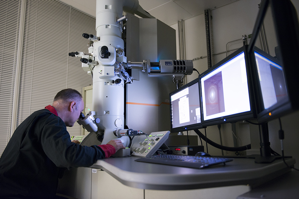 Photograph of a man looking into a microscope. He sits in front of several computer monitors. The device shown in the image is part of NNL's user access call.