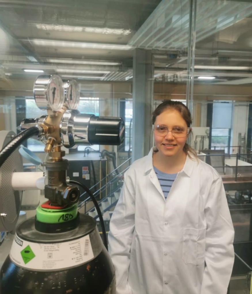 Photograph of Suzanne Jones in a white lab coat and goggles standing next to machinery in a lab.
