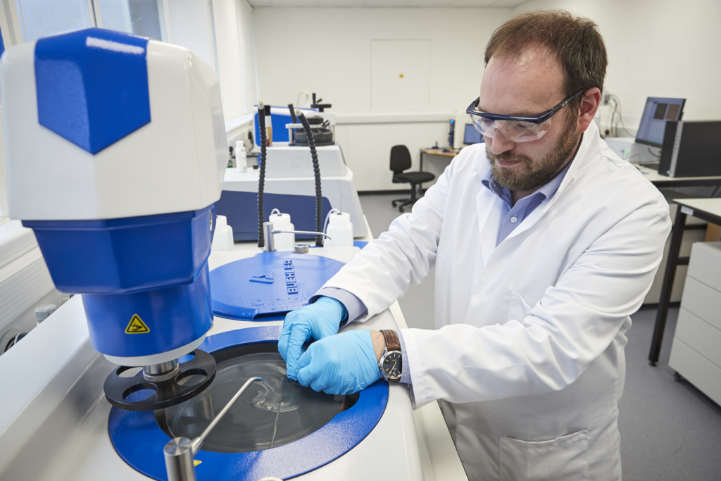 Photograph of Simon Middleburgh working in a lab. He wears safety goggles, a white lab coat and blue gloves while looking down and handling materials in front of some equipment on a table.