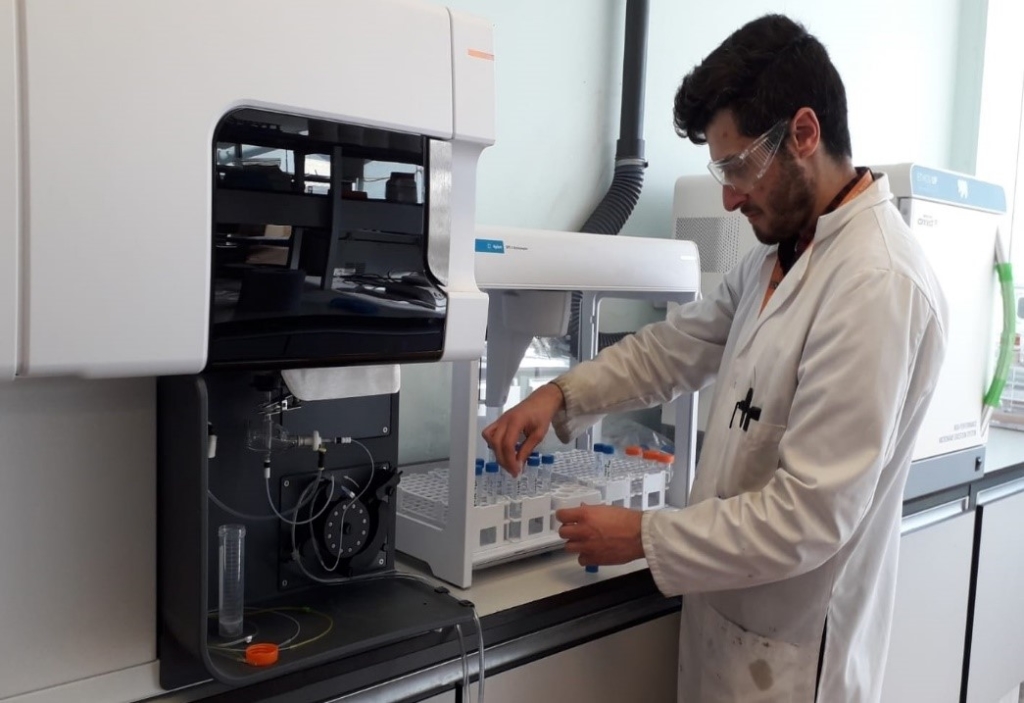 A photograph of AFCP researcher Antony Nearchou in a lab. He is wearing a white lab coat and facing equipment in a counter top in front of him.